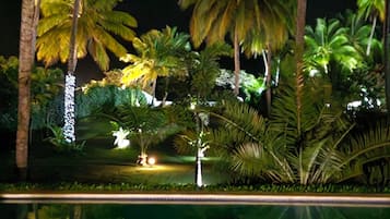 Vista noturna de um resort tropical, com palmeiras e piscina.