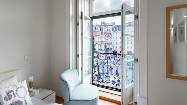 A bedroom with a bed, a chair, a nightstand, a window with a view of buildings, and a door. - The Restauradores Collection - Majestic (Lisboa)