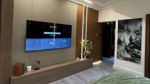 A modern hotel room with a large flat-screen TV mounted on the wall, a bedside table with a vase of flowers, a bed with a green throw, and a framed picture on the wall. - One Bedroom Luxury apartment in Maitama, Abuja (Abuja)