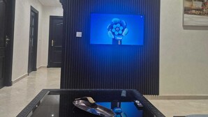 Um lobby moderno com uma televisão de tela plana grande fixada em uma parede escura, uma mesa de centro de vidro com um controle remoto, e uma porta que dá para outro cômodo.
