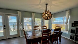 Uma área de refeições com mesa e cadeiras de madeira, grandes janelas com cortinas e um lustre.