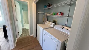 Uma área de lavanderia com duas máquinas de lavar e secadoras, uma prateleira com produtos de lavagem e uma porta que dá para outro cômodo.