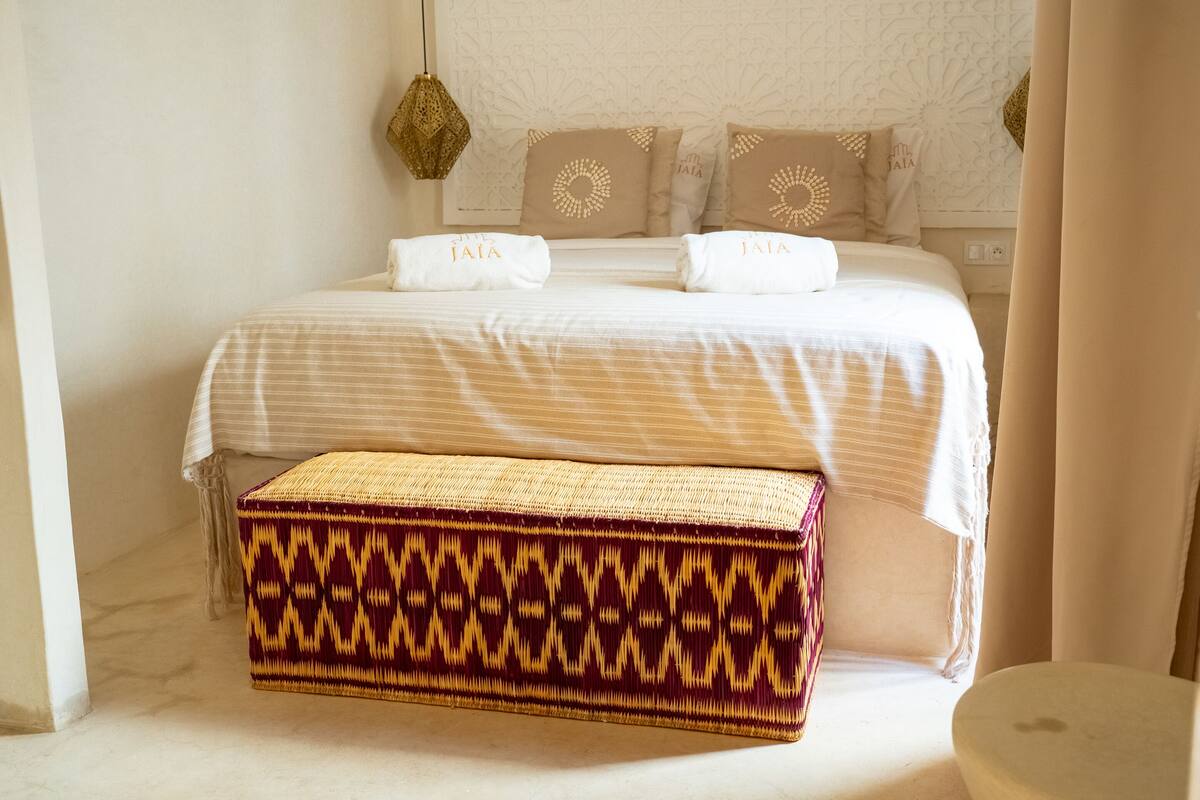 A neatly made bed with white linens and pillows, a woven bench, and a decorative wall panel.