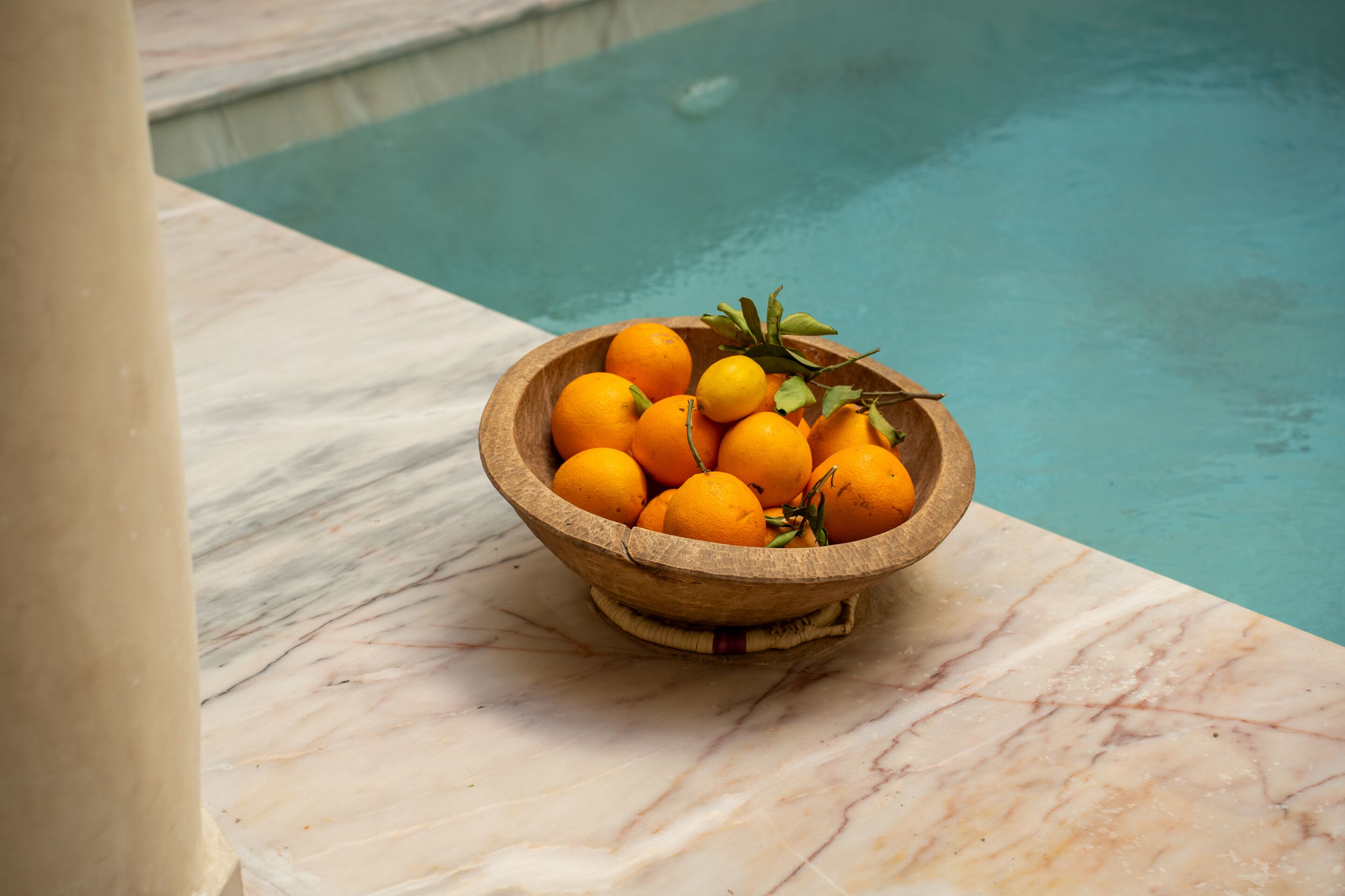 A wooden bowl of oranges on a marble surface near a pool.
