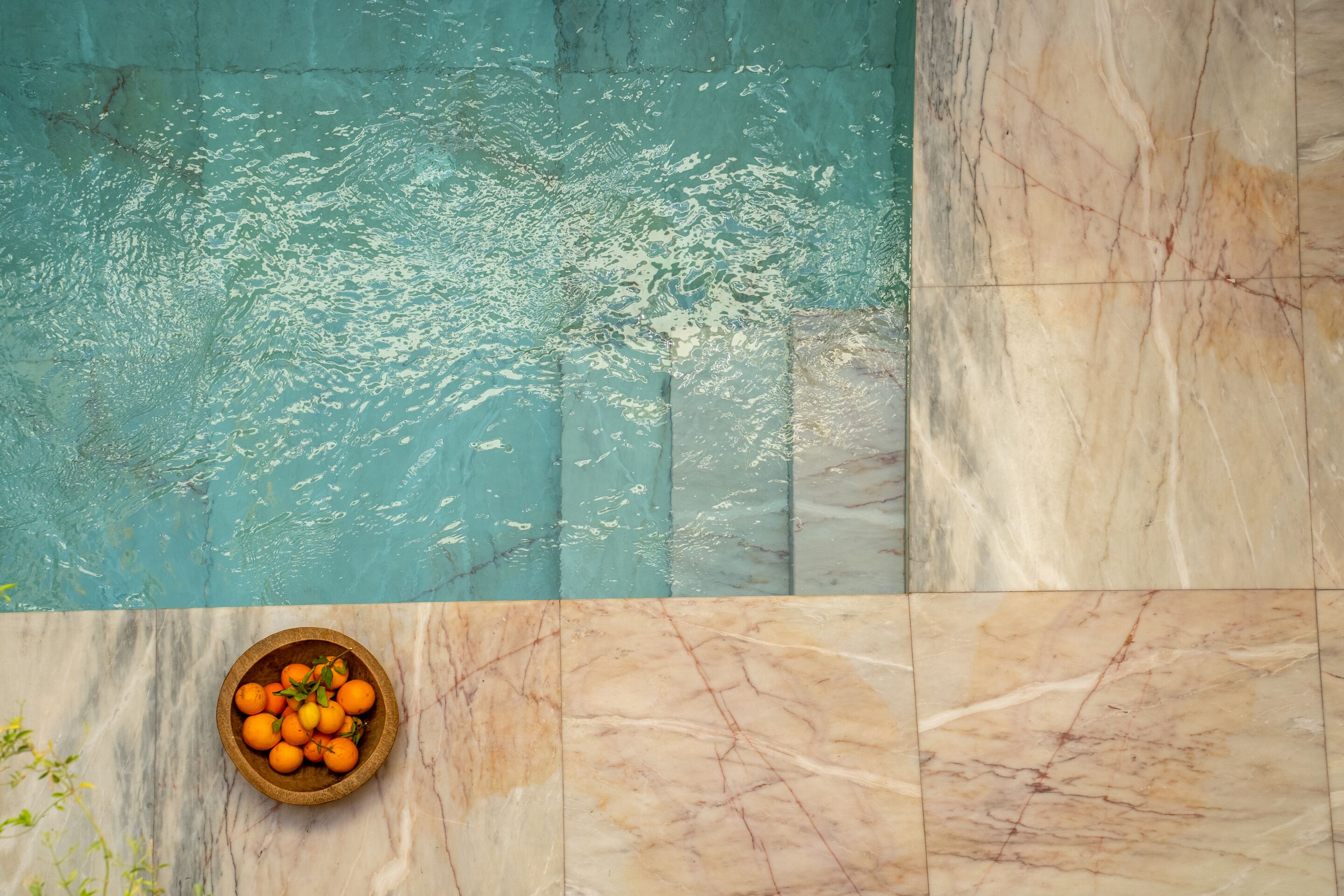 A pool with clear water, a bowl of oranges on a marble surface, and a tiled area with a marble pattern.