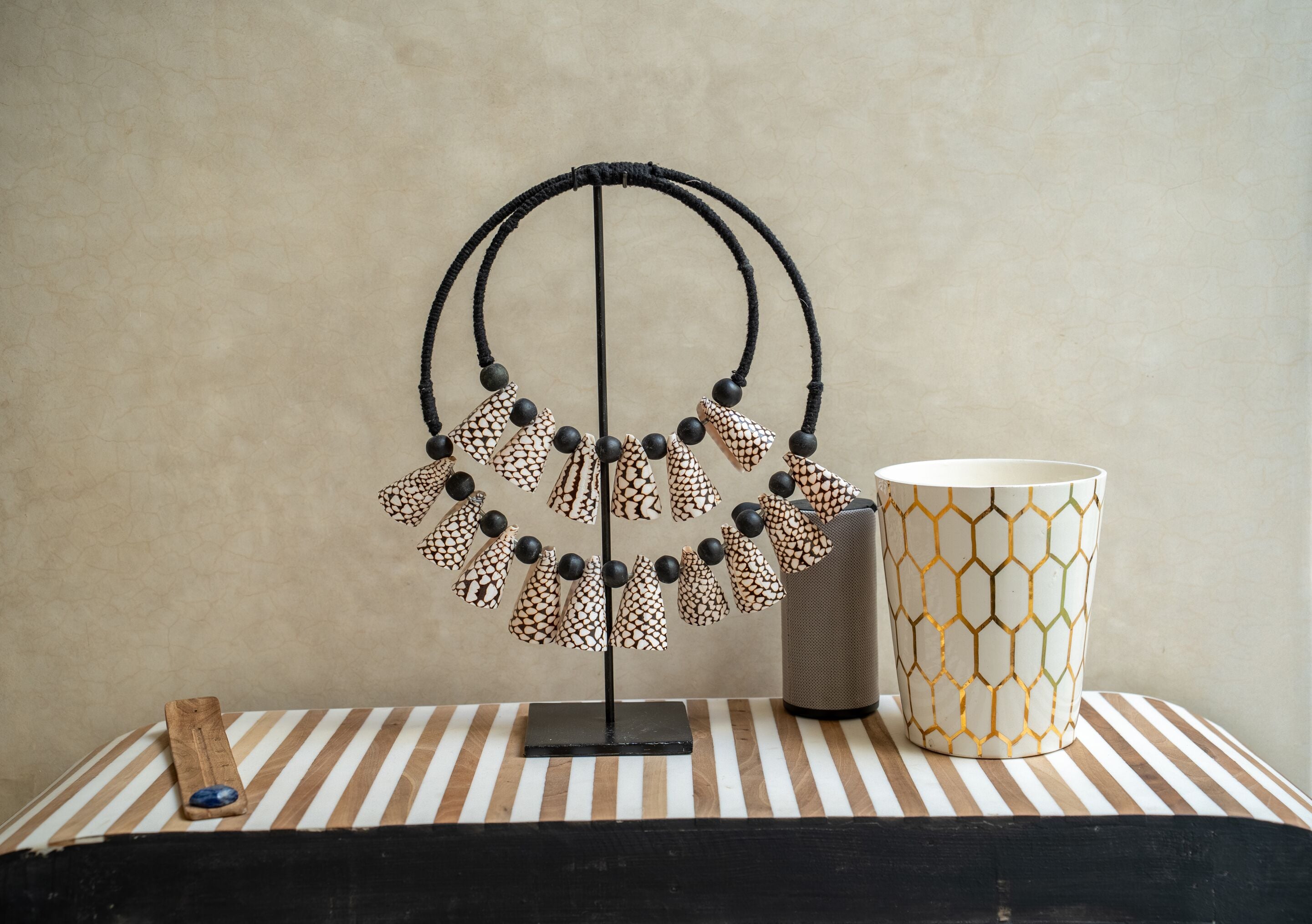 A display stand with a beaded necklace and a patterned vase on a striped table.