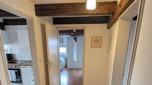 Un couloir avec un plancher en bois, des murs blancs et des poutres apparentes. Il y a une affiche sur le mur et une porte qui donne sur une autre pièce.