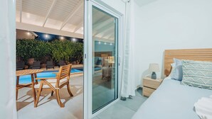 Ein Schlafzimmer mit einem großen Bett, einem Nachttisch und Blick auf einen Außenpoolbereich mit Tisch und Stühlen.