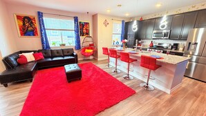 Un salon moderne avec un canapé en cuir noir, un pouf rouge et un tapis rouge. La cuisine est équipée d’appareils électroménagers en acier inoxydable et comprend un coin bar avec des tabourets rouges.
