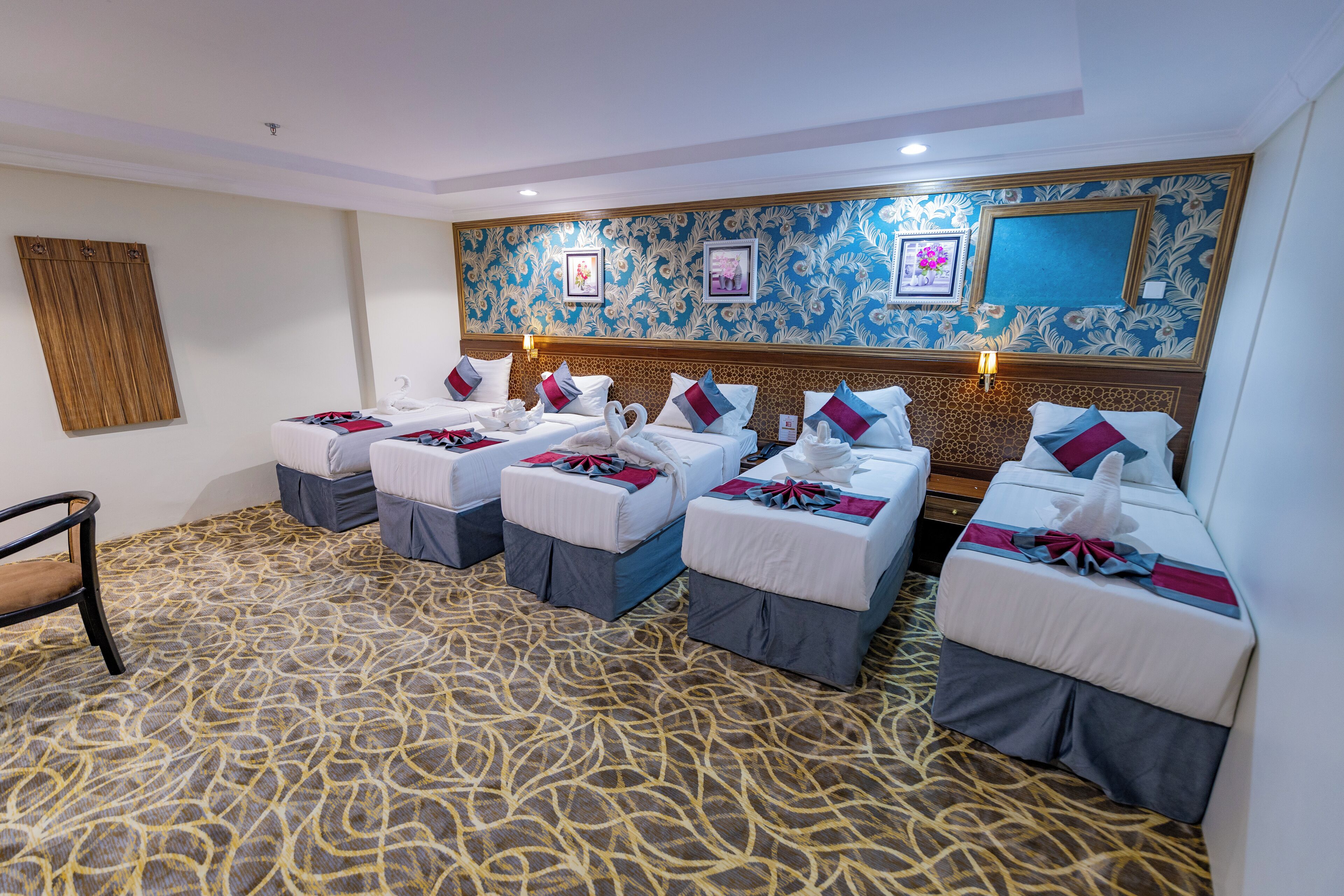 Ein Hotelzimmer mit vier Betten, einem hölzernen Kopfteil, einem gemusterten Teppich und Wandleuchten.