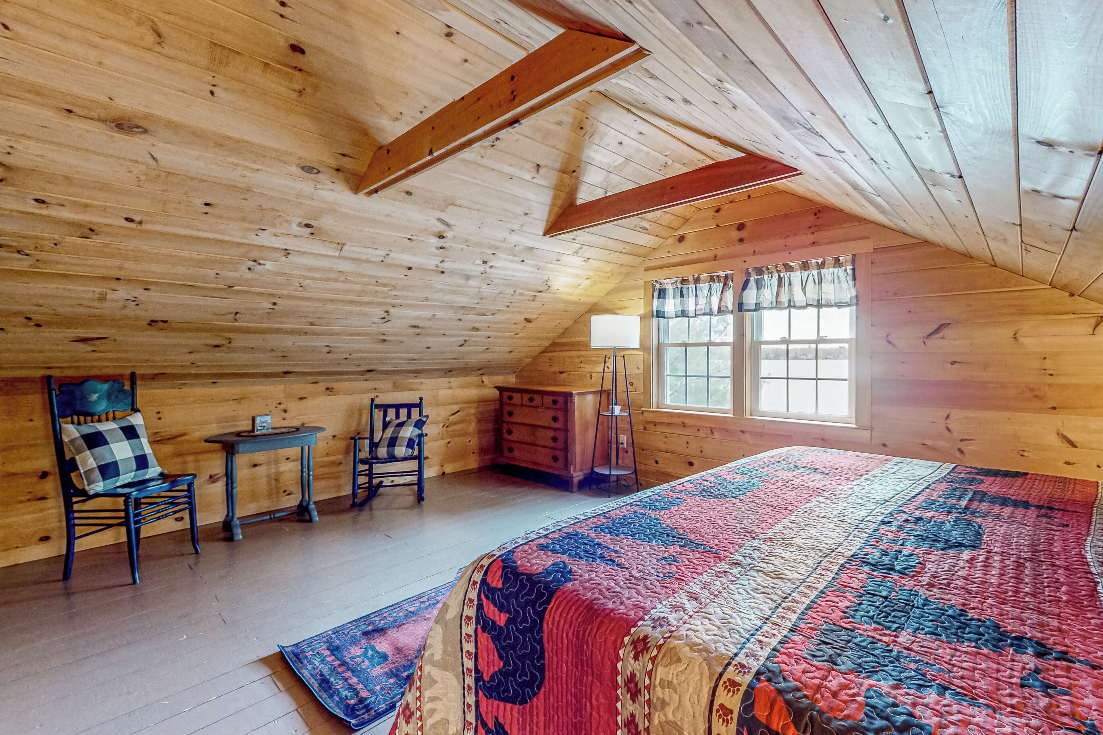 Un dormitorio acogedor en el ático, con una cama grande, paredes de madera, un ventanal con cortinas y una pequeña mesa con sillas.