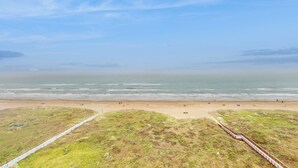 Ein Strand mit einer Holzpromenade, die zum Ufer führt, grasbewachsenen Dünen und einem klaren, blauen Himmel.