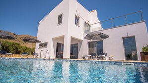 Un moderno edificio blanco con piscina, mobiliario exterior y montañas al fondo.