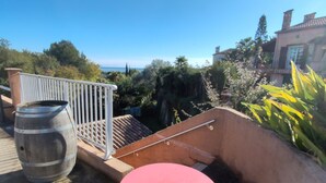 Une terrasse sur le toit, avec une table rouge et des chaises bleues, un grand tonneau en métal, et une vue sur la mer et la végétation environnante.