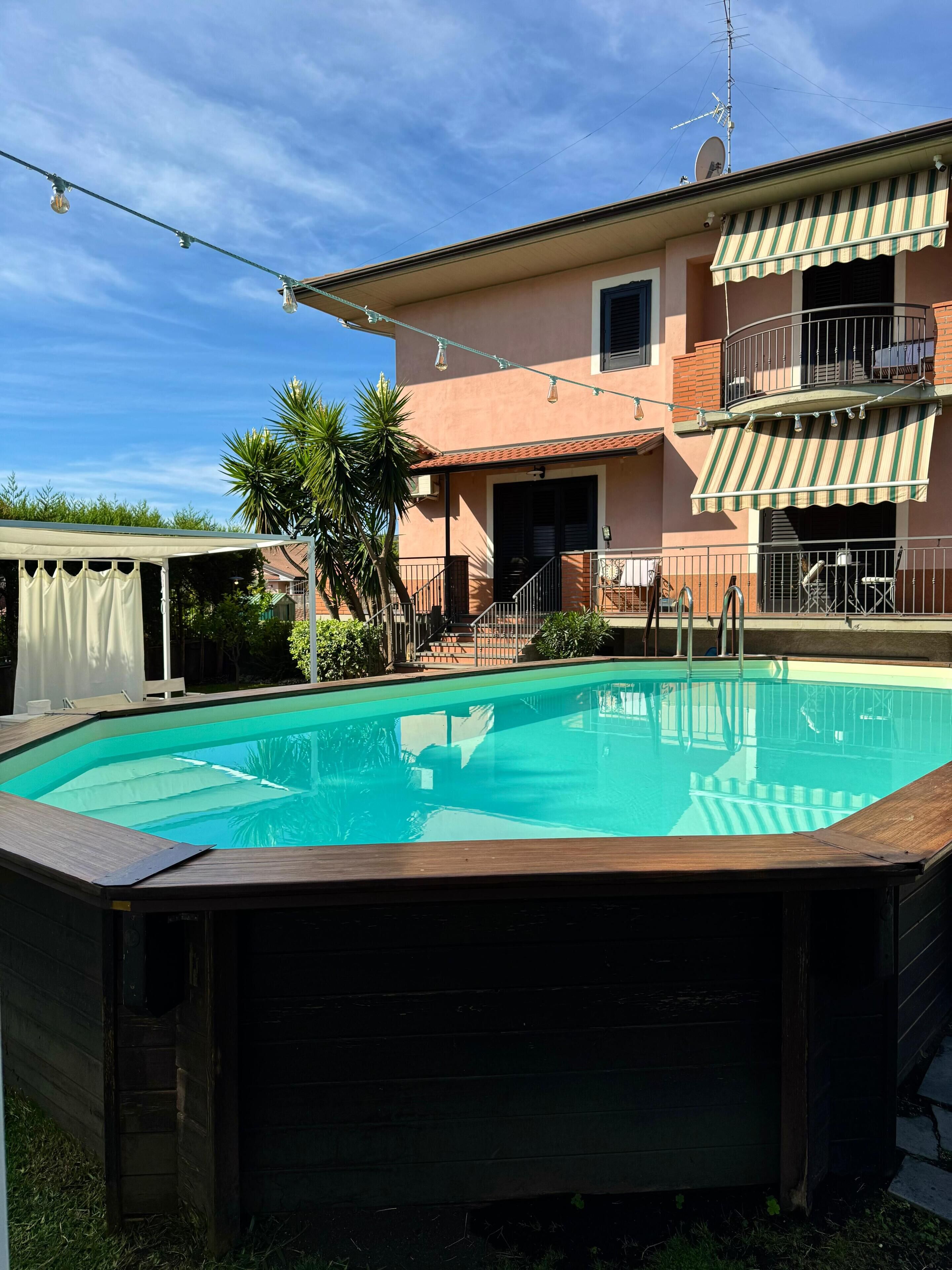 A swimming pool with a wooden deck, a covered patio area, and a two-story building with a balcony.