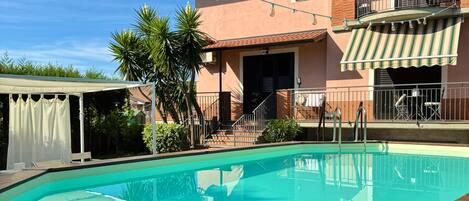 A swimming pool with a wooden deck, a covered patio area, and a two-story building with a balcony.
