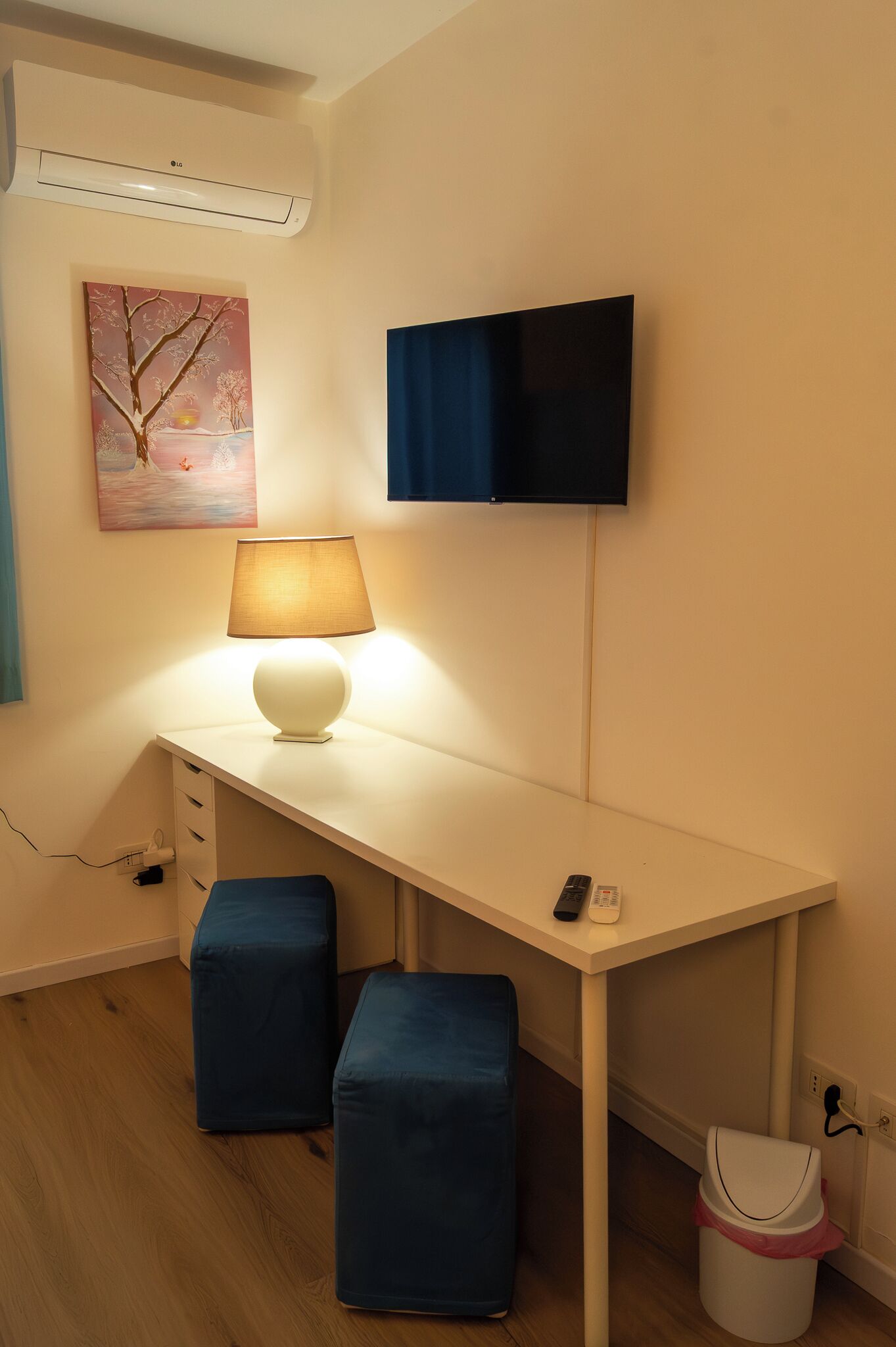 A small, neatly arranged room with a white desk, a lamp, a television, and a trash bin.