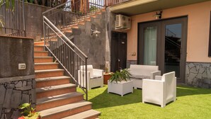 Ein Wohnhaus mit einer Treppe zu einem Balkon, einer Sitzgelegenheit im Freien mit weißen Korbmöbeln und einem gepflegten Rasen.