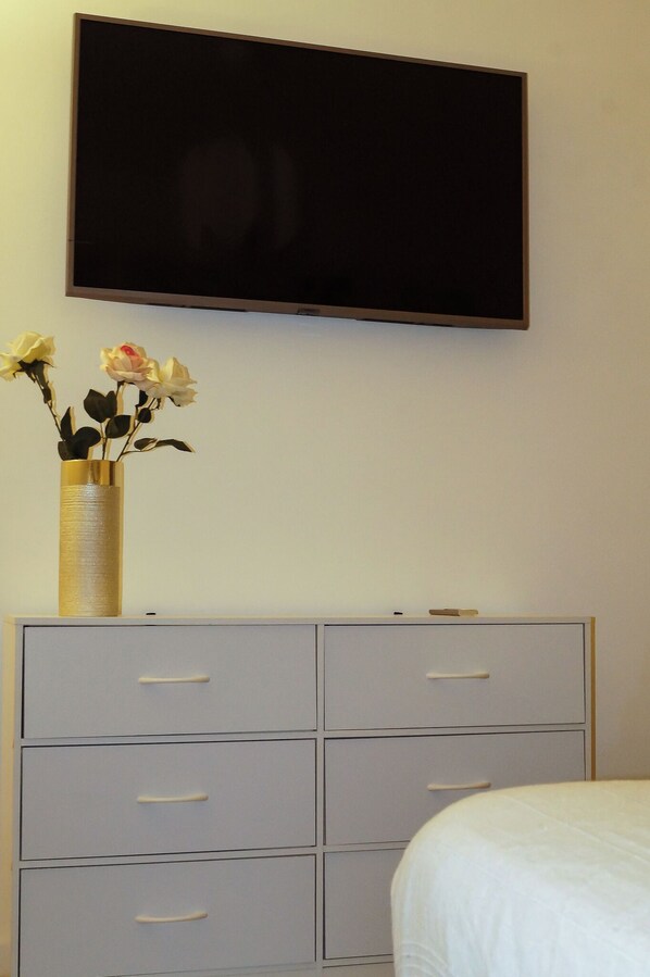 A bedroom with a bed, a dresser with drawers, a flat-screen TV mounted on the wall, and a vase with flowers on the dresser.
