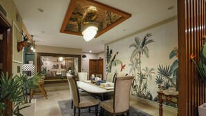 A dining area with a large table, chairs, and a chandelier. There are potted plants and a wall mural. - MG Luxury Stays (Panchkula)