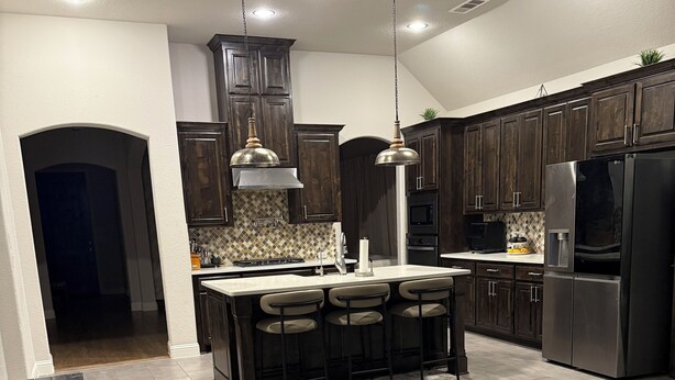 A modern kitchen with dark wood cabinets, a central island with bar stools, stainless steel appliances, and pendant lighting. - Spacious 3BR Home w/ Office & Backyard Vibes – Near Frisco. (Aubrey)