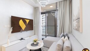 A modern living room with a large flat-screen TV, a sofa, a small round table with a vase of flowers, and a view of a building through a glass door. - Vogue | Aykon City Business Bay near Metro Station (Dubai)