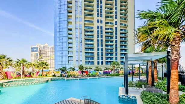 A swimming pool with a wooden deck, lounge chairs, and palm trees. - Vogue | Aykon City Business Bay near Metro Station (Dubai)