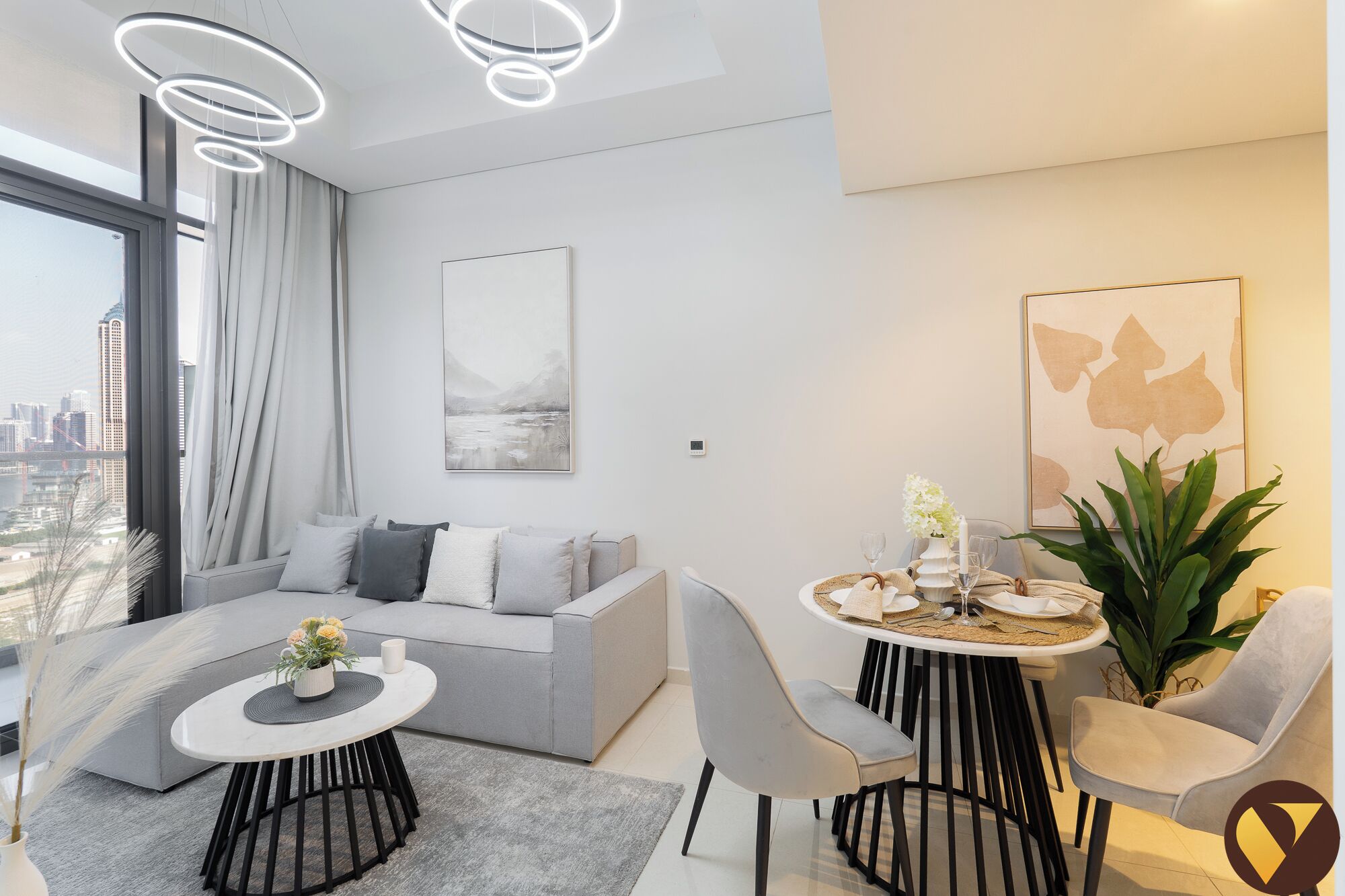 Una sala de estar moderna con un sofá gris, una mesa de comedor redonda con sillas, un ventanal con cortinas y vistas a la ciudad.