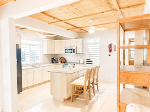 A modern kitchen with wooden ceiling beams, a central island with chairs, and a view of the outside through large windows.