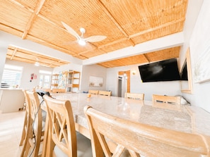 Une pièce spacieuse avec un grand plafond en bois, un ventilateur de plafond, une table à manger avec des chaises, un téléviseur fixé au mur et un coin cuisine avec des meubles en bois.