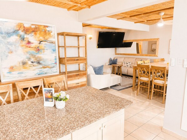 A modern living room with a TV, a painting, a dining area with wooden furniture, and a kitchen with a granite countertop.