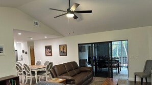 A modern living room with a brown leather sectional sofa, a dining area with white chairs, a ceiling fan, and a view of the outdoors through glass doors. - 3 bed/2 bath pool home & hot tub in Naples. Great neighborhood. 3 miles to beach (Naples)