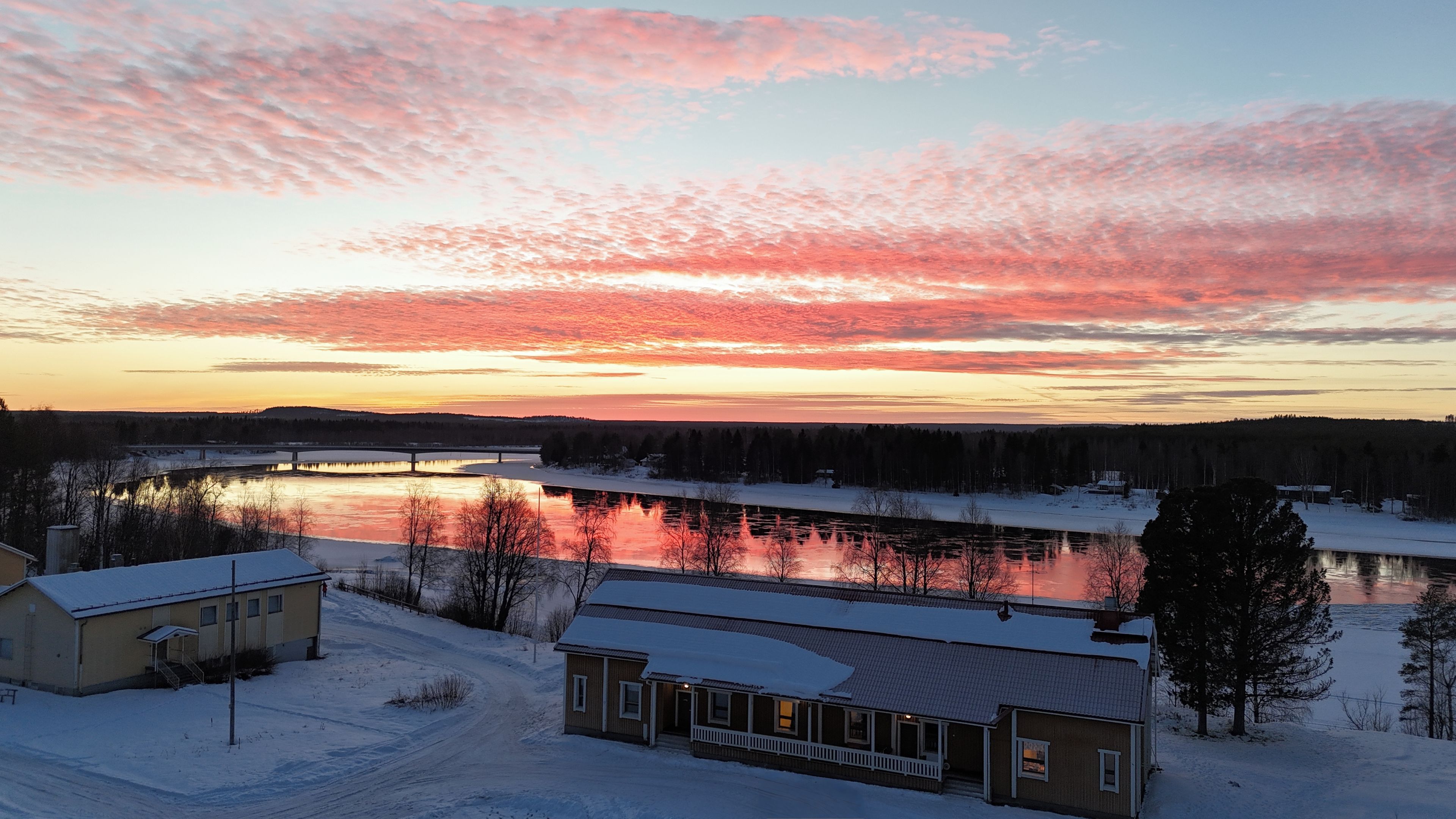 Talvinen maisema, jossa on joki, silta ja lumihunnun peittämiä rakennuksia.