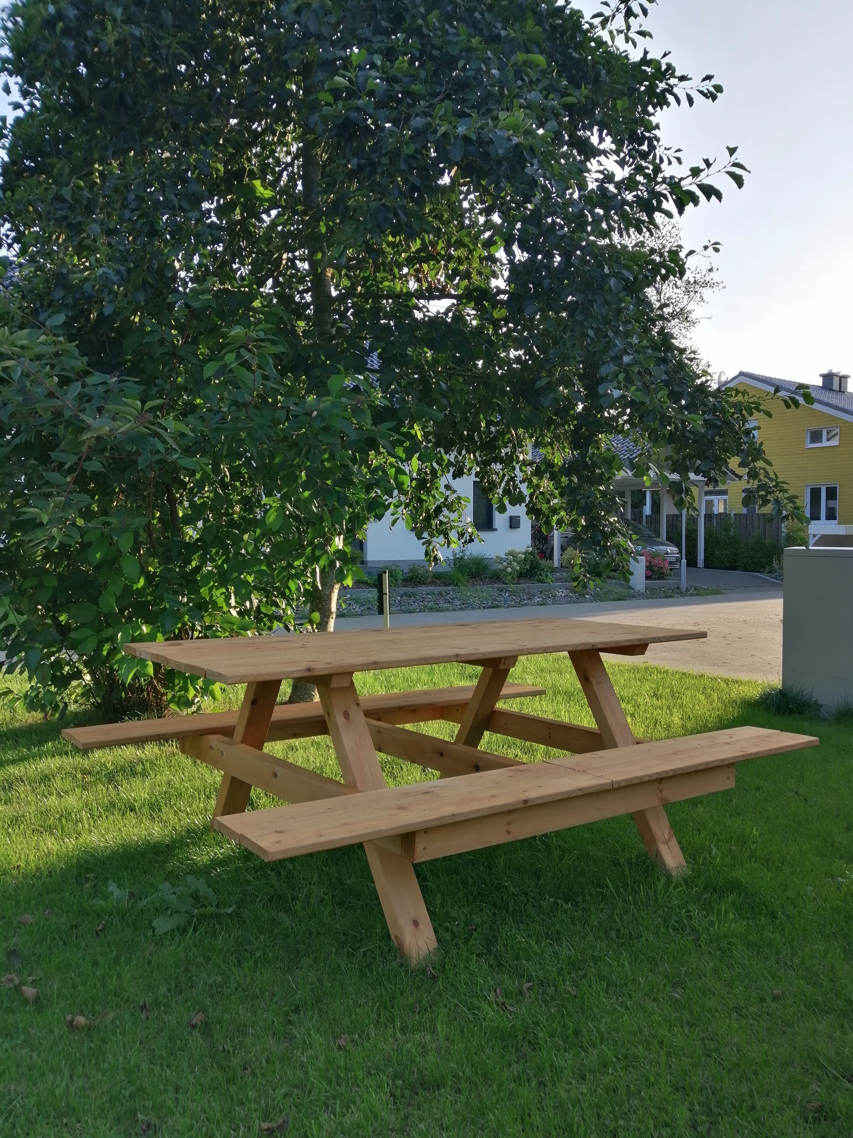 A wooden picnic table in a grassy area with a large tree and a building in the background.