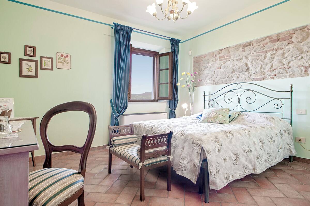 A bedroom with a bed, a chair, a desk, a window with curtains, and a stone wall.