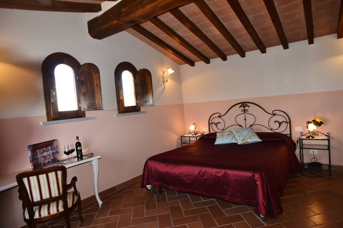 A bedroom with a large bed, a wooden chair, a small table with a wine bottle and glasses, and two wall-mounted lamps.