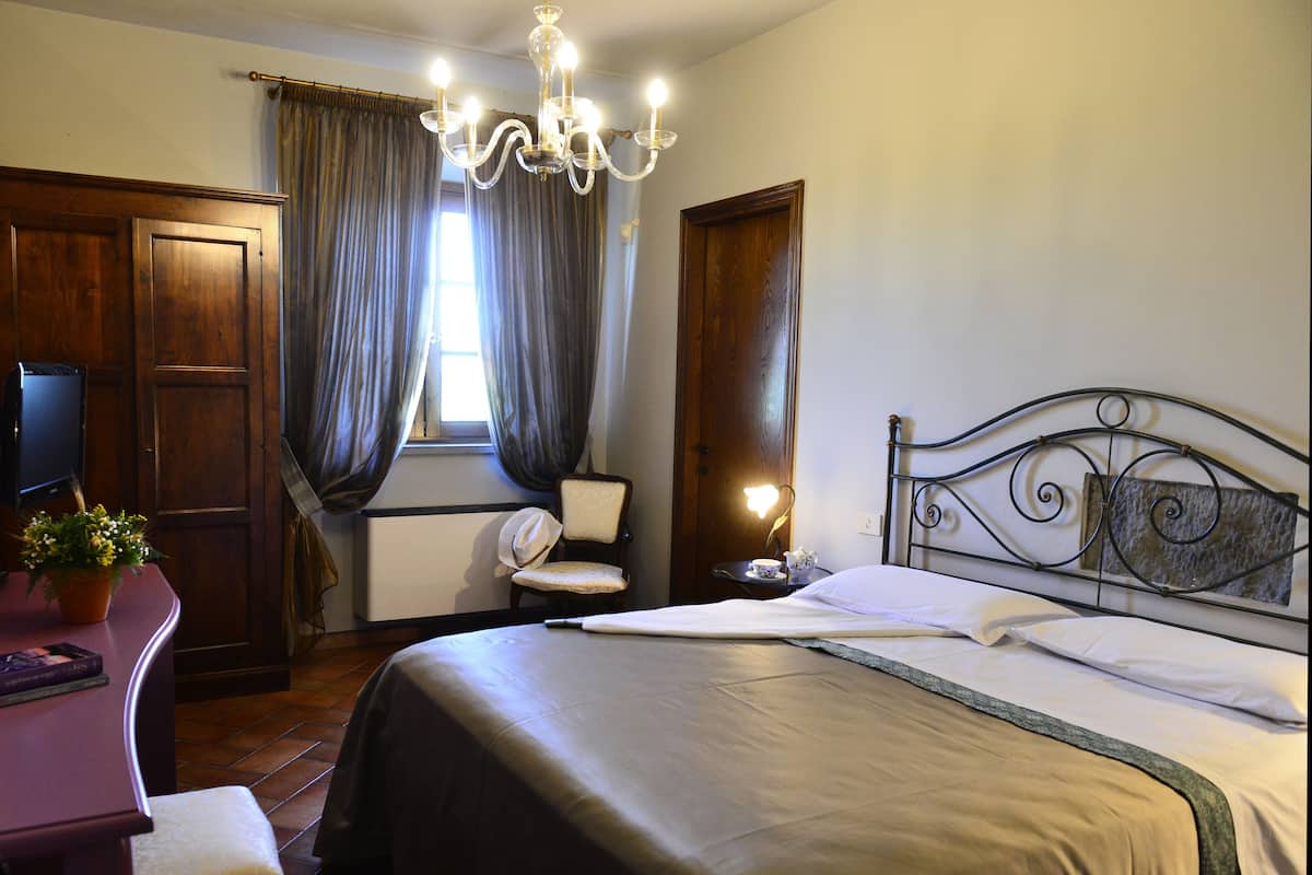 A bedroom with a bed, a desk with a computer, a chair, a TV, and a window with curtains.