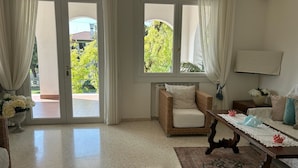 Una habitación amplia con un gran ventanal corredero que da a un espacio exterior, un sillón de mimbre, una mesa de madera con mantel de encaje y un aire acondicionado instalado en la pared.
