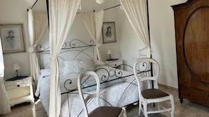 Una cama con dosel y cuatro columnas, ropa de cama blanca, un armario de madera y una silla blanca.