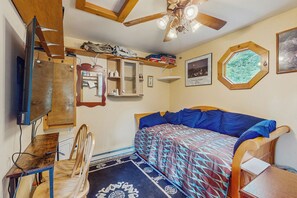 Una habitación compacta con cama, escritorio, silla, ventilador de techo y televisor en la pared.