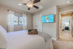 Ein Schlafzimmer mit einem Bett, einem Deckenventilator, einem Fenster mit Vorhängen, einer Tür und einem Fernseher an der Wand.