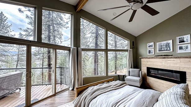 Una camera da letto con un letto grande, un ventilatore a soffitto, un camino e vista sulla foresta attraverso ampie finestre.