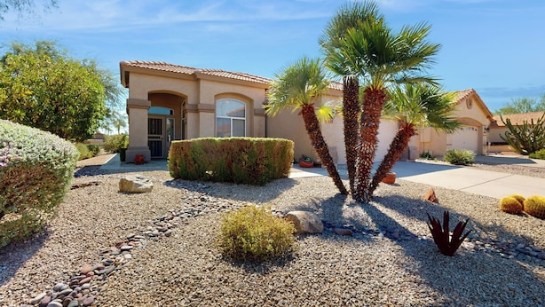 Una casa residenziale con un giardino curato, con una palma, arbusti diversi e un vialetto in ghiaia.
