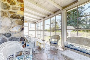 A covered patio with stone walls, wicker furniture, and large windows offering a view of trees. - Group Getaway on Working Farm! Near Saratoga (Ballston Lake)