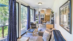 A cozy living room with a sofa, armchair, and coffee table. There is a dining area with a table and chairs, and a kitchen area with a microwave and sink. Large windows with curtains provide natural light and a view of the outdoors. - Walk to Cafes! Storybook-Style Fairbanks Studio (Fairbanks)