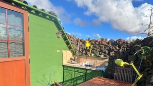 Uma pequena construção com telhado verde, mesa e cadeiras de madeira, um abajur amarelo e um muro de pedra ao fundo.