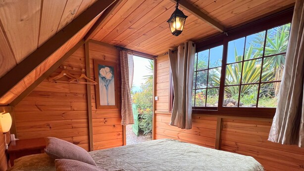 Quarto de cabana de madeira com cama, almofadas, um quadro e janela com cortinas.