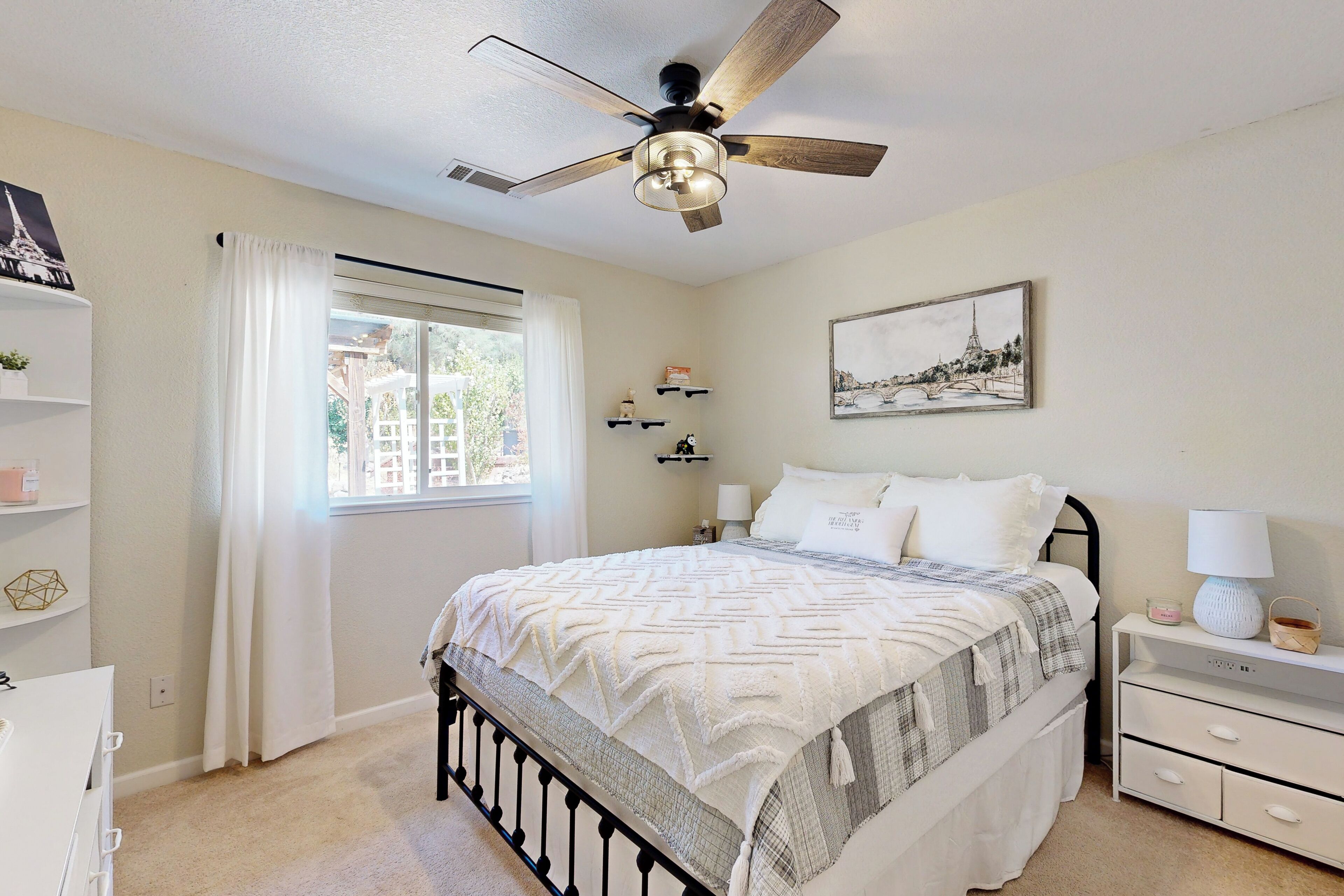A bedroom with a bed, bedside tables, a ceiling fan, a window with curtains, and wall-mounted shelves.