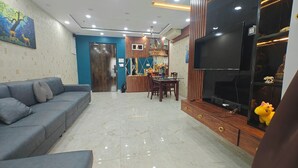 A modern living room with a grey sofa, a dining table with chairs, a TV on a wooden stand, and a kitchen in the background. - 3bhk fully furnished highrise apartment in a luxerious complex with lake view (Kolkata)