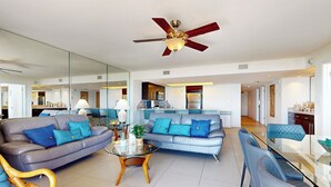 Ein modernes Wohnzimmer mit einem Glastisch, zwei blauen Sofas, einem Deckenventilator und einer Küche im Hintergrund.